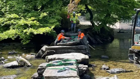 Workers in high-vis vests and hard hats use a digger to move huge slabs of stone crossing a shallow ford. Trees line the river bank. 