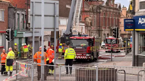 Emergency service personnel, wearing yellow or orange hi-vis jackets, on a town centre road which has been cordoned off due to a fire. Several fire engines can be seen parked in a road and a ladder on one of the engines has been extended upwards.