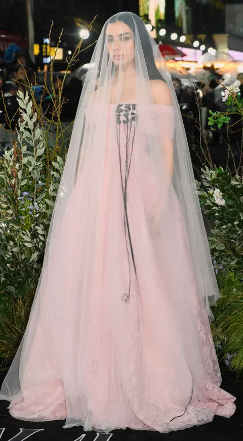 Getty Images Charli XCX stands posing for a picture on the red carpet wearing a light pink floor-length dress with matching sheer veil that reaches the floor. 