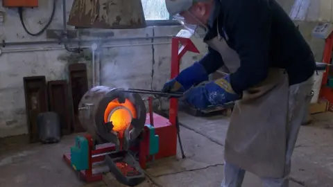 A man with a poker attending to a furnace that is full of fire and molten tin glowing bright orange. He is wearing big blue gloves, a face visor and a brown apron.