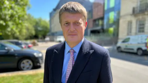 Conservative Richard Clewer looks at the camera - he wears a dark suit jacket, blue shirt and pink tie. Blurred behind him is the Wiltshire Council headquarters on a sunny day with a few cars parked outside.