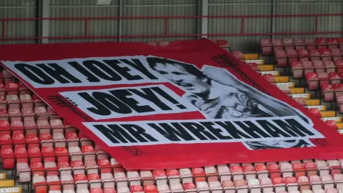 A large red banner spread across a block of red plastic seats in a football stadium. The banner reads in large black font "Oh Joey Joey! Mr Wrexham" with a large black and white photo of Joey Jones on the right hand side of the text. 