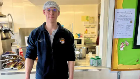 Project lead Reuben Mardell-Kearns standing in the kitchen. Packets of food can be seen behind him. He is wearing a white hair net and glasses. He is looking at the camera.
