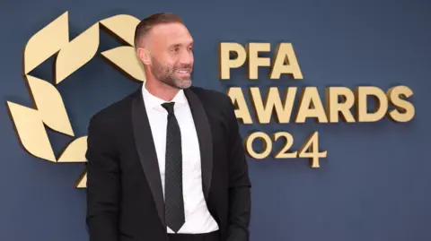 Calum Best is stood in a suit and tie wearing a white shirt. He is smiling to the right stood in front of a dark blue wall with gold writing and a logo on it. The words say PFA AWARDS 2024. 