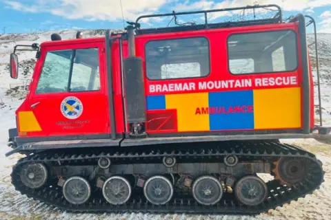 Red vehicle with tank-style caterpillar tracks, red body with white writing which says Braemar Mountain Rescue, and the word ambulance in red on blue and yellow checks, on snow, with snowy hillside in background.