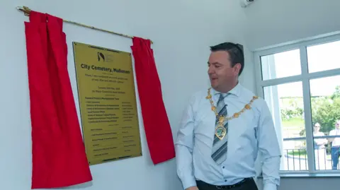 DCSDC Mayor Ruairí McHugh is wearing his mayoral chains and a blue shirt and striped tie. He is standing beside a golden plaque that has two 