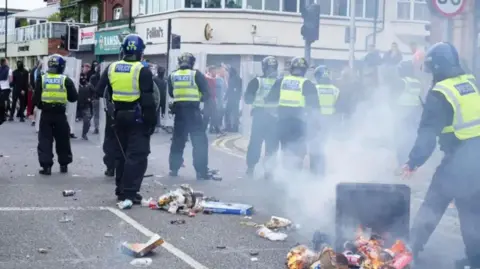 Back view of group of helmeted police officers on a street, to the bottom right a bin is on its side with burning rubbish and smoke rising above it
