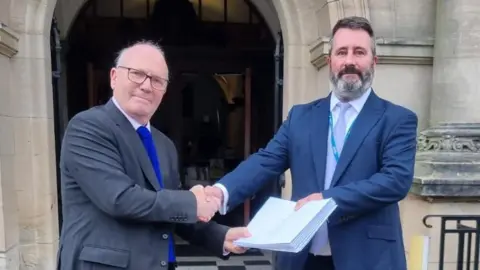 Chris Hall, from SHOUT, hands the petition to Daniel Harry, North Yorkshire Council's head of democratic services. Two men, both wearing suits, light-coloured shirts and tie, shake hands and exchange a paper folder.