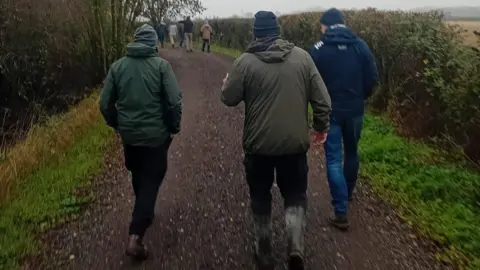 James Clements A group of people are walking on a narrow path, which has hedges and trees either side. There are three men walking away from the camera and a group ahead of them.