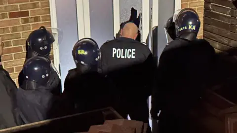 BBC / Oliver Whitfield-Miočić Kent Police officers wearing riot uniforms, helmets and vests stand outside a home with a pane of broken glass from the front door visible following a forced entry into the property.