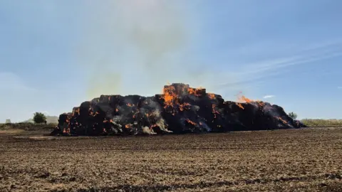 A large pile of hay piles burns under a blue sky. The hay is blackened by the fire, with orange flames and dark smoke rising above it. 
