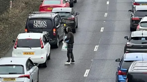 Eddie Mitchell A man approaches a white car, holding some water and crisps. 