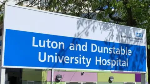 Bedfordshire Hospitals NHS Foundation Trust A close up of the Luton and Dunstable hospital sign. A tree casts a shadow on the sign. 