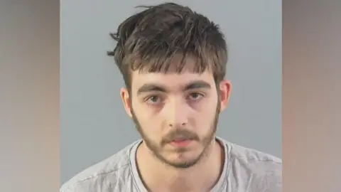 Carlo Tritta, who is wearing a grey jumper, is staring directly at the camera in this custody mugshot. He has straight, light brown hair and a thin beard.
