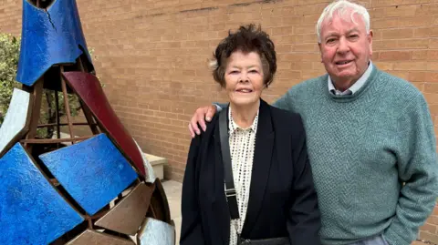 Gavin Kermack / BBC An elderly couple is standing next to the piece of art from the above picture. The woman has short dark hair and is wearing a white blouse patterned with coloured spots under a dark blazer. The man has short white hair and is wearing a green jumper over a collared shirt. He has his arm around her.
