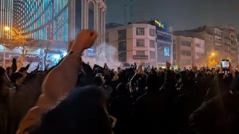 Getty Images Hundreds of people stand in the streets of Tehran with their fists raised in the air. There is a small amount of smoke hovering above their heads in parts of the picture.