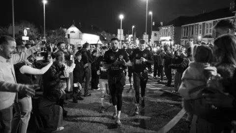 Jekyll Photography A black and white photo at night of the friends running along a residential road towards the camera, with large crowds gathered alongside, filming on cameras