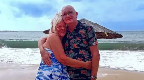 Facebook An image of a blonde haired woman in a blue dress hugging a man in a floral shirt on a beach.