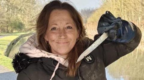 Gill Shilvock holding a knife that she has pulled out of a canal, with the waterway in the background