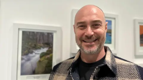 Adam is smiling stood infront of some paintings. He is wearing a black and cream shirt/jack with a black top underneath.