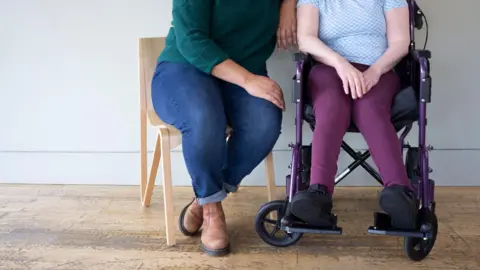 Two people sitting close together, one is a woman in jeans and a green jumper sitting next to a women in a wheelchair. You can only see their lower bodies, not their faces.