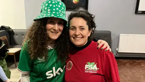 Dr Penny Miles Two women smiling wearing Ireland and Wales colours. Dr Penny Miles is wearing a red football shirt and has her dark, curly hair tied back. A woman wearing an Ireland shirt and a green bucket hat covered in clovers has her arm around Dr Miles.