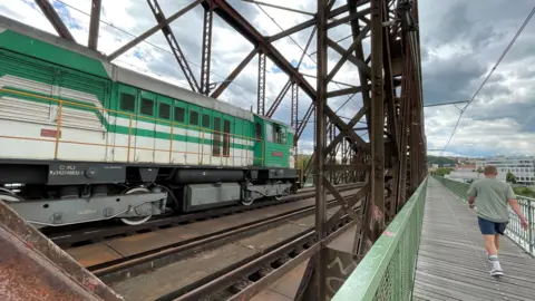 A green engine is seen on a rusty metal bridge, as a man walks alongside on a pedestrian walkway.
