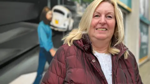Emma Baugh/BBC Debbie, a woman with blonde hair, wearing a burgundy puffer jacket, smiling for the camera. 