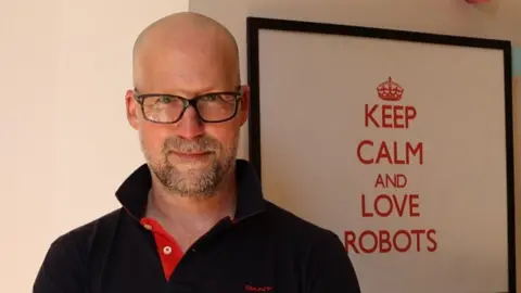 Ingmar Posner Ingmar Posner in glasses and a black polo shirt stands in front of a pictures that reads: "Keep calm and love robots."