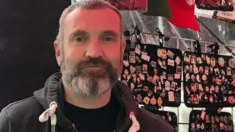 Robbie MacDonald Market stall holder Mike Gallavan on his stall standing in front of displays of badges. He has short grey hair and a beard and is wearing a black hoodie.