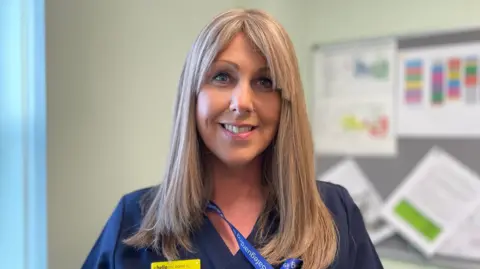 Teresa Cope has long blonde hair with a parted fringe. She is smiling, wearing navy scrubs and standing in an office with a noticeboard on the wall.