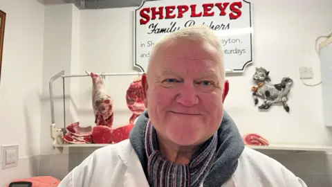 Richard is wearing a white coat over the top of a winter jacket and scarf. Behind him there's meat hanging up ready to be sliced up, and a sign that reads 'Shepley's Family Butchers.'