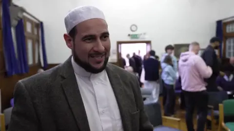 BBC Shaykh Adam Kelwick wears a Taqiyah, white shirt and grey blazer as she speaks to the right of the camera, there are crowds of people behind him in a room