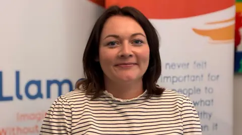 Yvonne photographed indoors in front of promotional display banners, wearing a striped long-sleeved top.
Yvonne has shoulder length brown hair and is smiling at the camera.