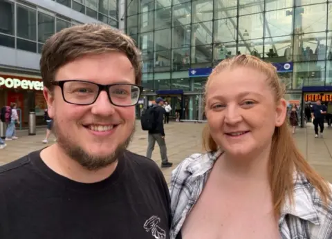 Michelle Adamson/BBC Shannon and Luke Harrison. Shannon has long blonde hair and wears a white lumberjack shirt with black tartan. Luke has brown hair and a beard with spectacles. He wears a black T-shirt. They stand outside Manchester Piccadilly Station. 