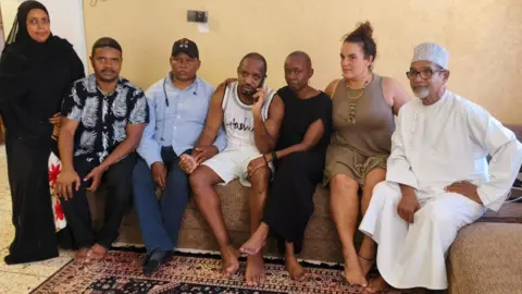 KNCHR/X Boniface Mwangi (centre in a white T-shirt and shorts, his wife Njeri (in black dress) and others seated on a coach in a room