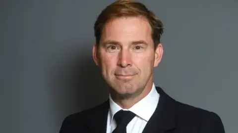A man wearing a black jacket and tie and mainly white shirt is prominent in the photo. He is looking at the camera and standing in front of a plain grey background.