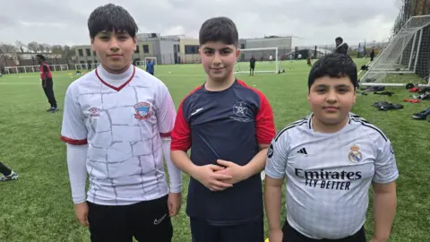 BBC Three young boys with dark hair are standing side-by-side on a football pitch, smiling. They are all wearing football shirts. Behind them are football coaches wearing bibs, and goal posts.