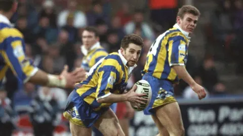Getty Images Several men in yellow, blue and white rugby jerseys, with one man in the centre holding a Puma branded, white rugby ball. The man in the centre has medium length brown hair and long yellow socks that nearly reach his knees