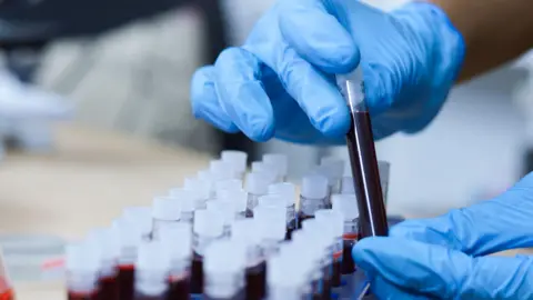 A test tube containing a dark red liquid is being held by two hands wearing blue plastic gloves.