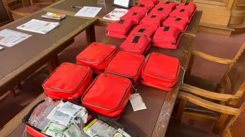 More than 20 bleed kits, packaged small red zipped bags, lined up on a brown wooden desk