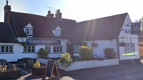 The Three Horseshoes is an old building with a tiled roof and white walls. There are signs showing its name, and benches and tables at the front of the building