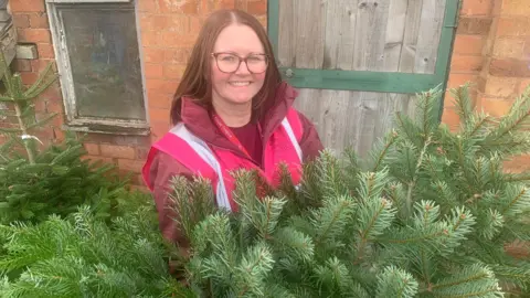 Katharine House Hospice A woman smiling in a pink vest over a burgundy jacket stands with a Christmas tree outside a red-brick building. She has loose shoulder-length dark hair and wears glasses.