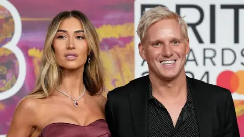 Jamie Laing and Sophie Habboo on the Brit Awards red carpet in 2024. Jamie wears a black shirt with small white spots and a black jacket over it, while Sophie wears a read dress without straps. She has long blonde hair and is slightly looking past the camera, while Jamie has short blonde hair and is smiling at the camera..