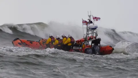 Helen Cowan An orange lifeboat heads out to sea among large choppy waves. It is an open top boat with inflatable sides and there are four crew aboard each wearing a yellow drysuit, orange lifejacket and white helmet