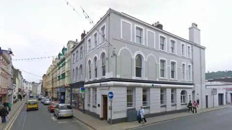 Google View of the Ramsey Lloyds Bank building on the corner of Parliament Street and Christian Street. It is a large three-storey building in grey and white that has rows of windows on each level, some of which are arched at the top on the second floor. There are cars travelling and parked on Parliament Street in front of it and people can be seen walking past the side of the building on Christian Street.