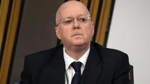 PA Media Close-up of Peter Murrell wearing glasses, a dark suit, white shirt, and tie, seated indoors in a Scottish Parliament committee room against a backdrop of curtains, looking slightly upward.
