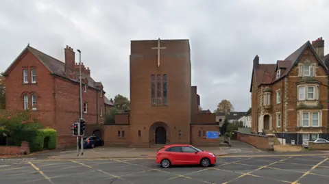 A modern brown-bricked church with a large cross at the top of the building. There are three stained-glass windowed panes at the front of the building. The church is on a main road, which has a red car travelling down it. 