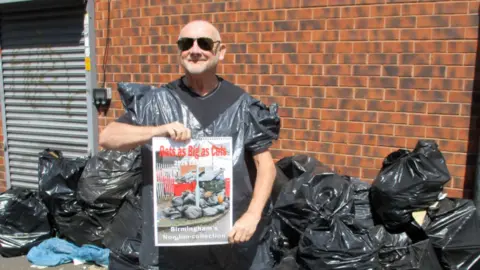 Kevin is bald and wearing black sunglasses, he is wearing a black t-shirt with a bin bag fashioned into a top, over the top of it. He is holding a calendar that says "rats as big as cats". In the background behind him there is a pile of black bin bag with rubbish scattered on the pavement