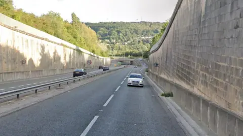 Image of a dual-carriageway with a number of cars driving on it. In the background can be seen trees and a roundabout. On ether side of the road are stone wall embankments. 